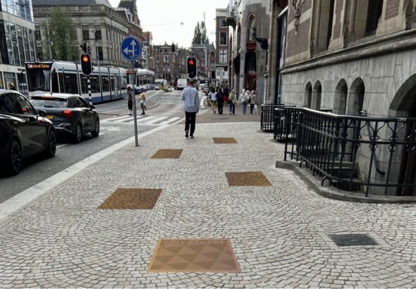 Op de foto is een trottoir naast een Amsterdamse straat te zien. Tussen de tegels van het troittoir zijn diverse koperkleurige tegels te zien, die deel moeten uitmaken van de Walk of Pride
