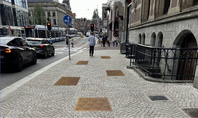 Op de foto is een trottoir naast een Amsterdamse straat te zien. Tussen de tegels van het troittoir zijn diverse koperkleurige tegels te zien, die deel moeten uitmaken van de Walk of Pride