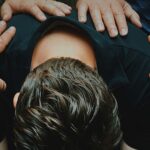 Op de foto is een man te zien, voorovergeboven zittend op een stoel. Zijn gezicht is niet te zien, we zien de achterkant van zijn schouders en zijn achterhoofd. Op de schouders en de rug van de man zijn verschillende handen te zien.
