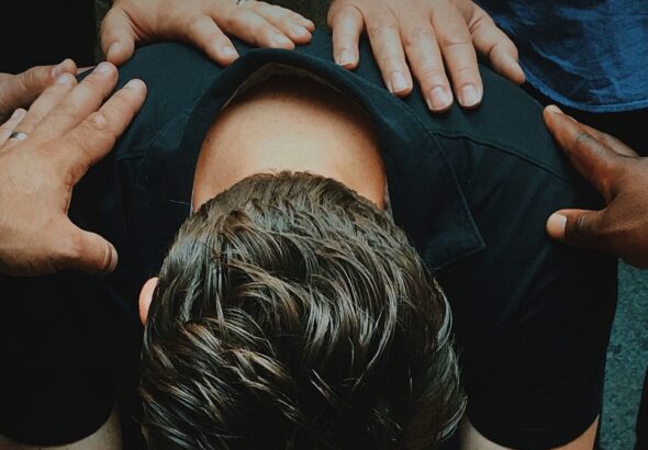 Op de foto is een man te zien, voorovergeboven zittend op een stoel. Zijn gezicht is niet te zien, we zien de achterkant van zijn schouders en zijn achterhoofd. Op de schouders en de rug van de man zijn verschillende handen te zien.