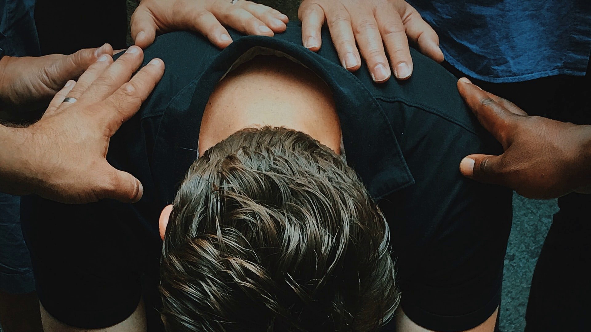 Op de foto is een man te zien, voorovergeboven zittend op een stoel. Zijn gezicht is niet te zien, we zien de achterkant van zijn schouders en zijn achterhoofd. Op de schouders en de rug van de man zijn verschillende handen te zien.